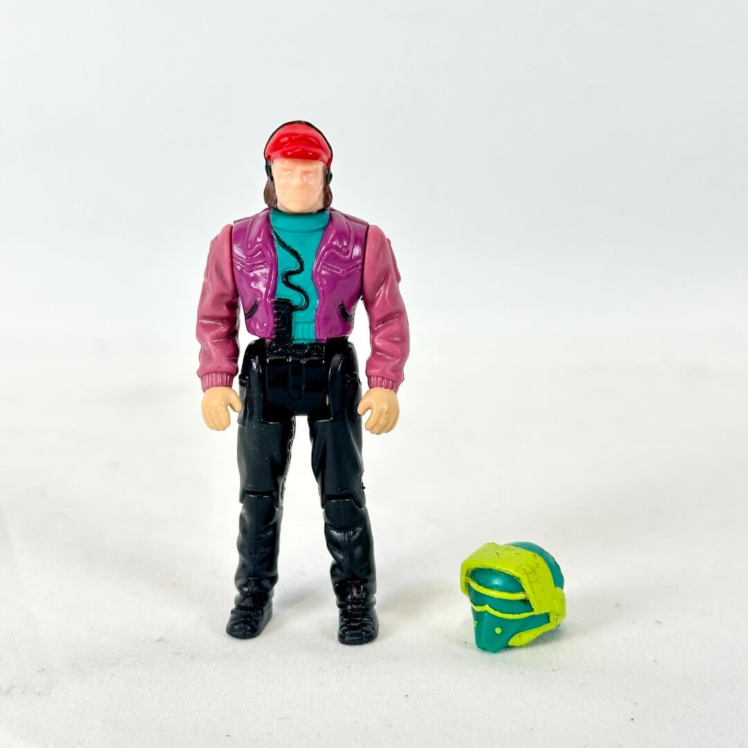 Action figure wearing a red cap, magenta jacket, teal shirt, and black pants, standing next to a green and yellow helmet.