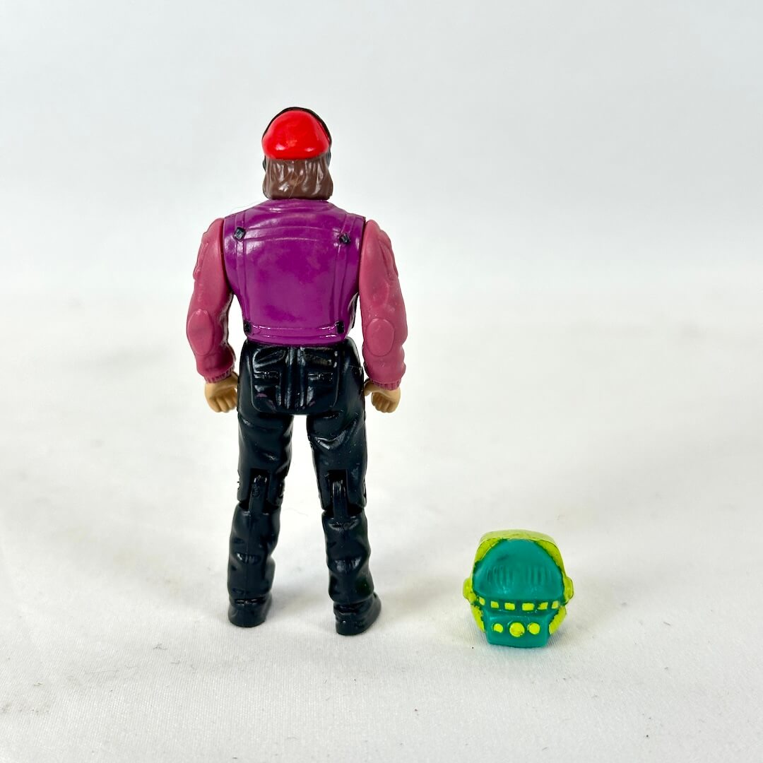 Action figure wearing a red cap, magenta jacket, teal shirt, and black pants, standing next to a green and yellow helmet.