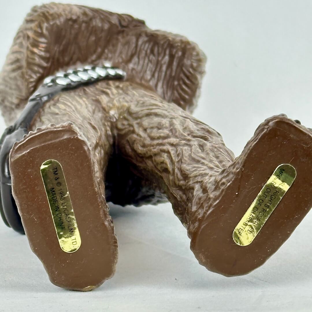 Close-up of a figure with textured brown surface and gold labels on bottoms of feet on a white background