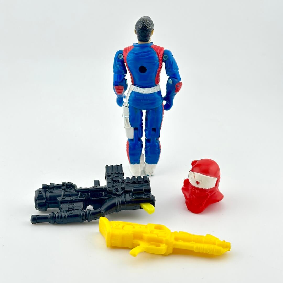 GI Joe action figure in blue flight suit with  multicolored accessories photographed on a white background. 