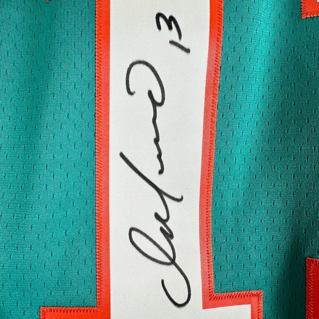 Close-up of a teal mesh sports jersey with a white number outlined in orange stitching, featuring a black handwritten signature.