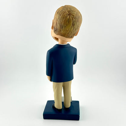 Bobblehead figure of a person wearing a dark blue shirt and beige pants on a white background
