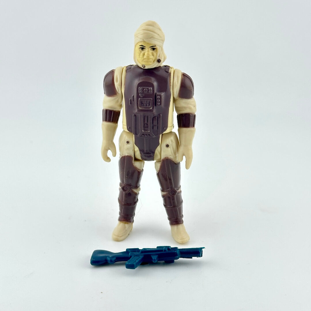 A vintage plastic action figure of a sci-fi soldier with beige skin and dark brown armor, standing next to a blue blaster.