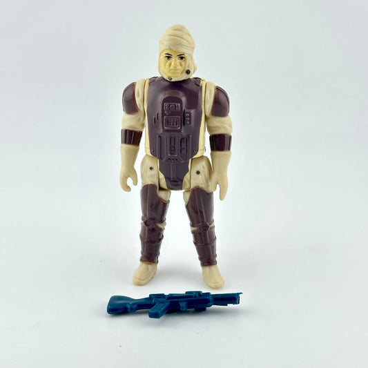 A vintage plastic action figure of a sci-fi soldier with beige skin and dark brown armor, standing next to a blue blaster.