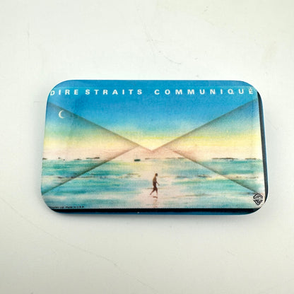 A rectangular metal pin with rounded corners, featuring the band Dire Straits' 'Communique' album artwork.
