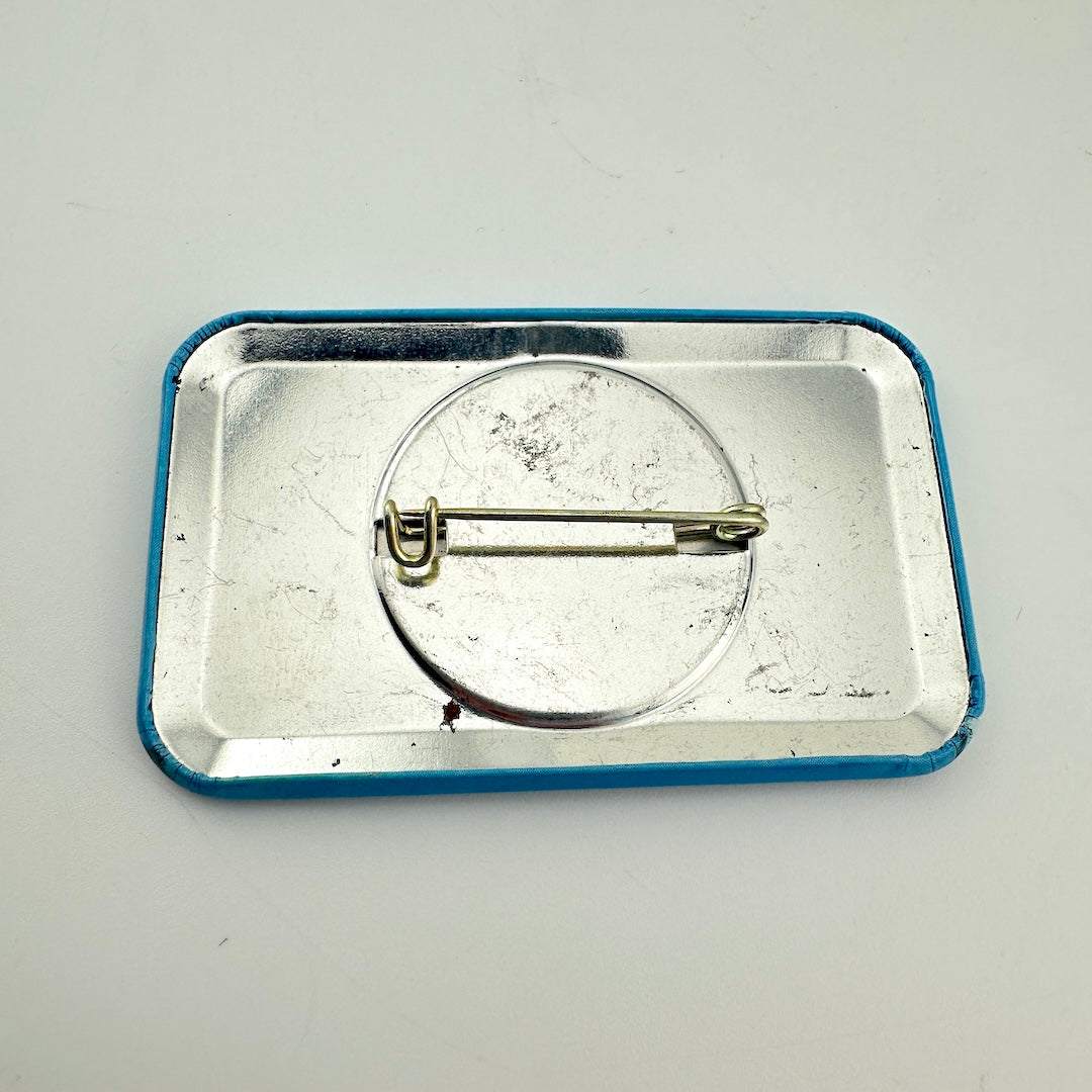 The back view of a rectangular pin badge with a blue border & a silver metal pin fastener.