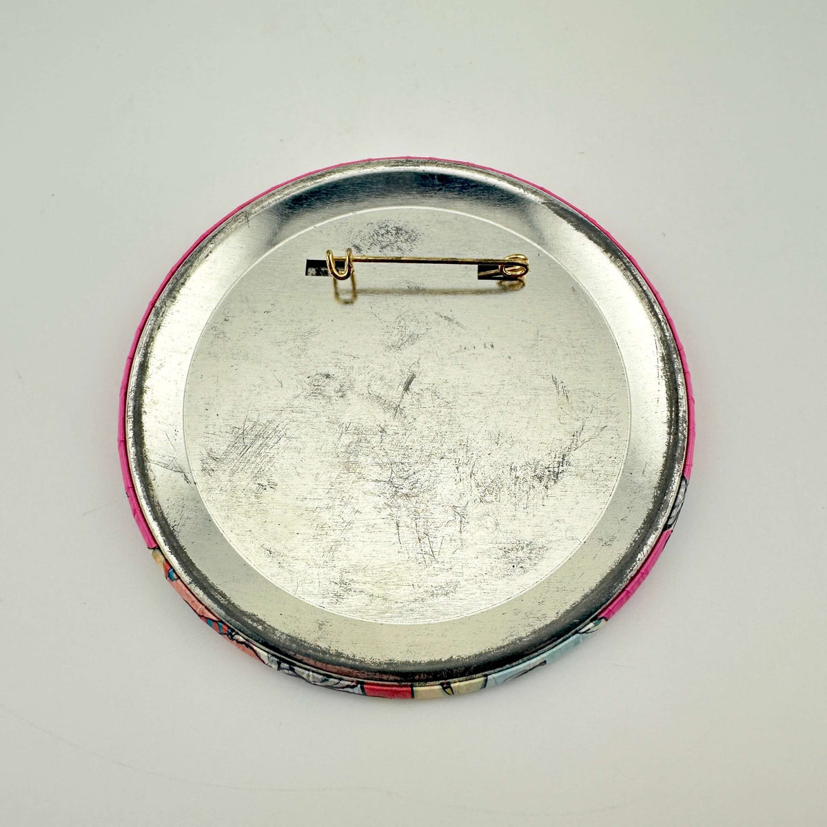 The back of a round pinback button badge, showing a silver metal backing, a gold safety pin clasp.