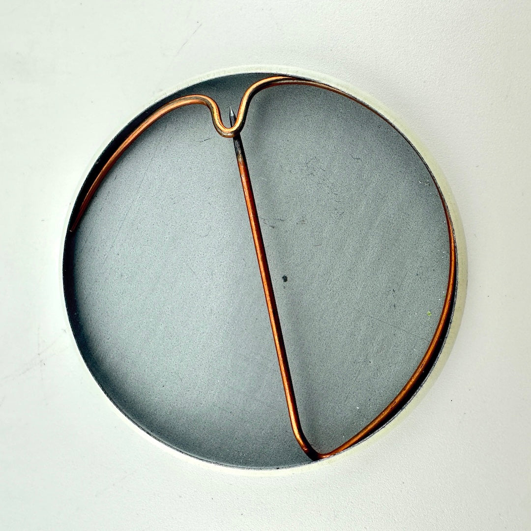 A circular gray pin with a copper-colored wire frame or loop running through the center.
