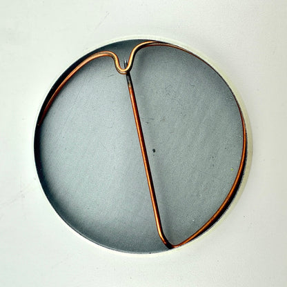 A circular gray pin with a copper-colored wire frame or loop running through the center.