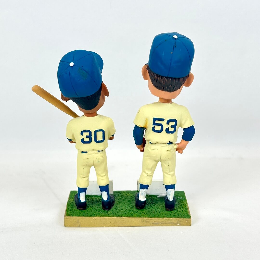 A pair of vintage bobblehead baseball figures shown from the back, standing on a green grass base. 