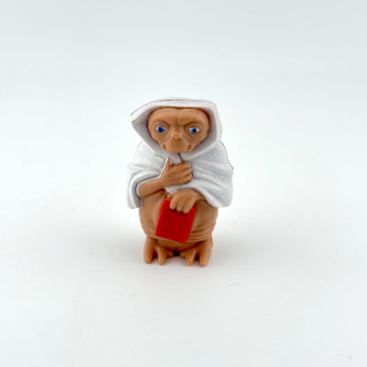 Small toy of E.T. in blanket holding red rectangle on white background. 