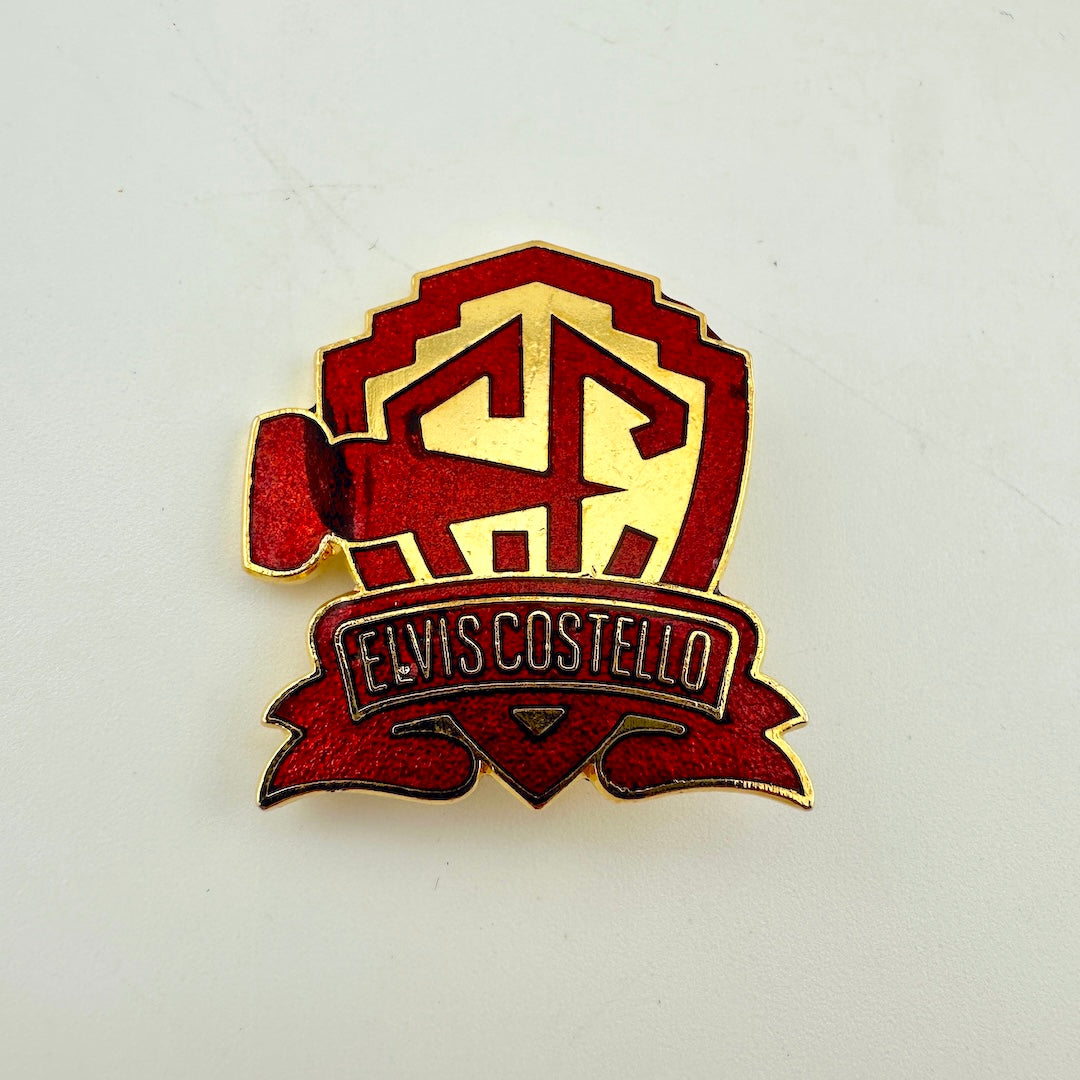 A red and gold Elvis Costello enamel pin featuring a shield with the initials 'EC' and a banner reading 'ELVIS COSTELLO' 