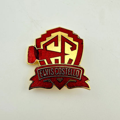 A red and gold Elvis Costello enamel pin featuring a shield with the initials 'EC' and a banner reading 'ELVIS COSTELLO' 