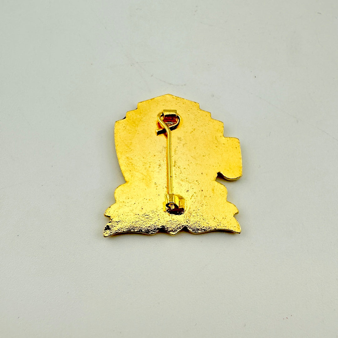 A gold-colored enamel pin shown from the back, featuring a stylized silhouette with a safety pin clasp.