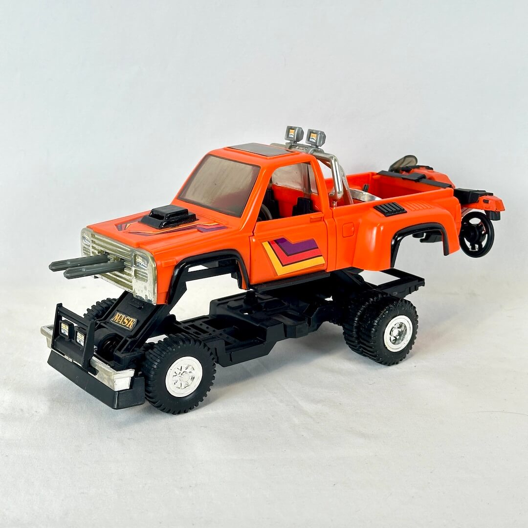 An orange and black toy pickup truck with chrome-like silver details, photographed on a plain light gray studio background.