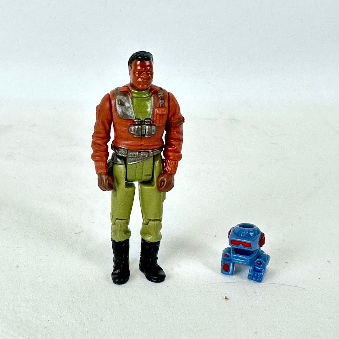 A vintage action figure of a man in an orange jacket, green pants, and black boots, standing next to a helmet.