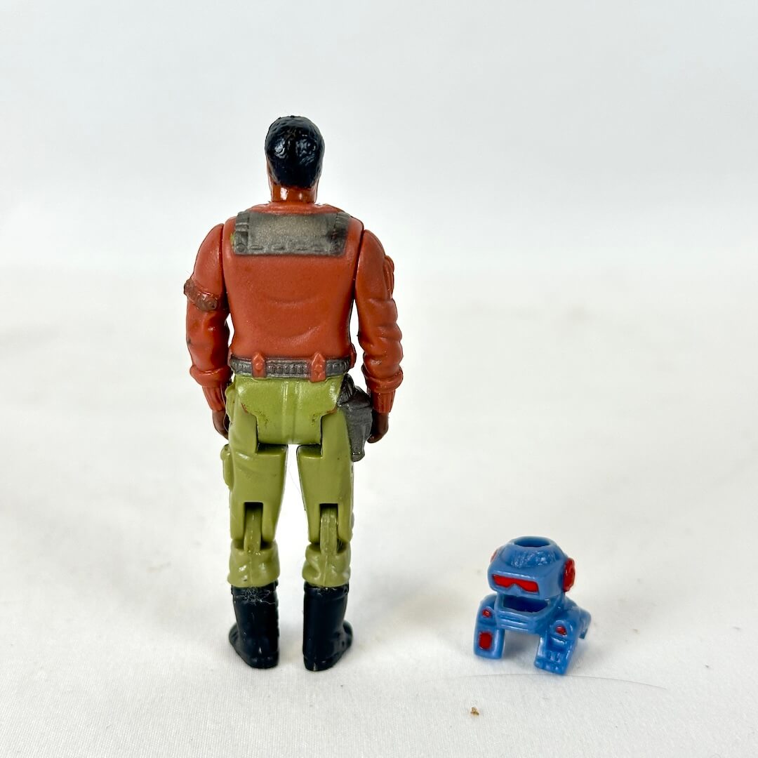 A vintage action figure shown from the back standing next to helmet accessory. 