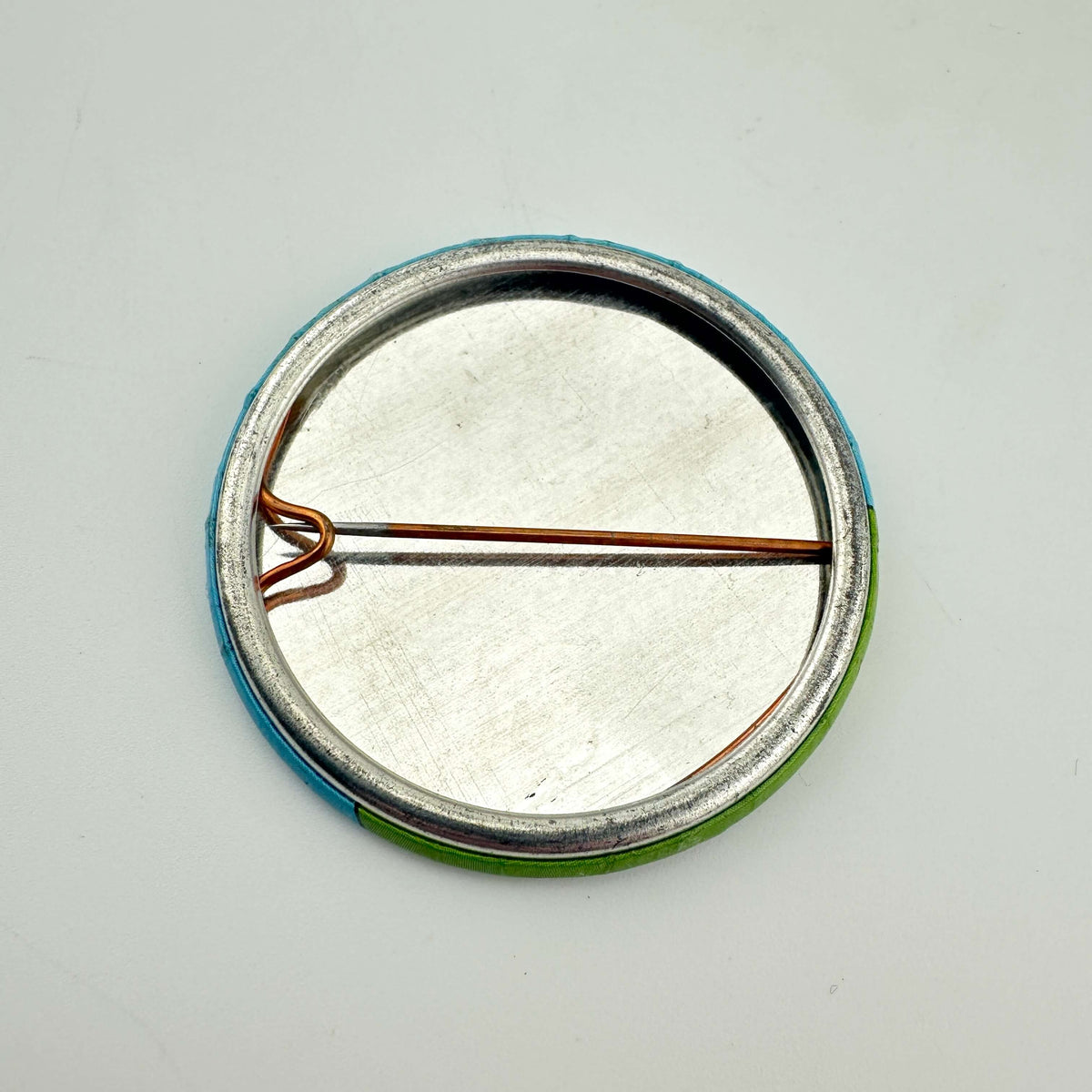 The back of a round pinback button badge showing a silver metal pin loop and backing with a blue and green fabric rim.