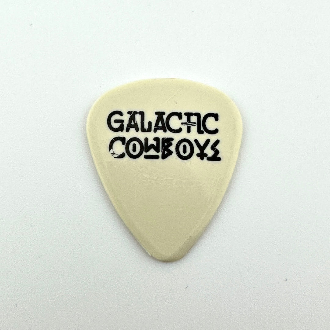 A cream-colored guitar pick with the words 'GALACTIC COWBOYS' printed in a black stylized font in the center.