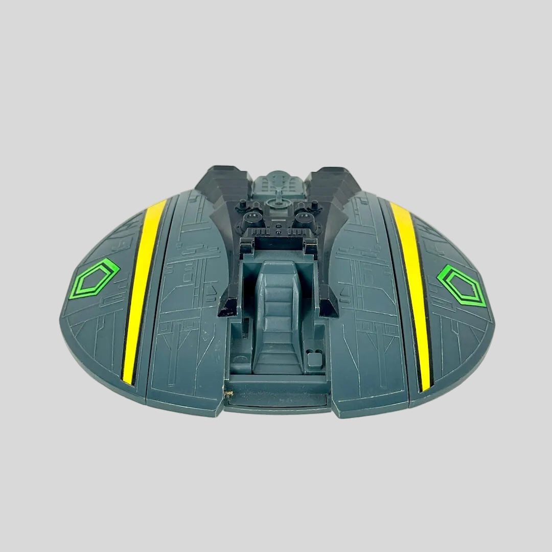 A toy spaceship accessory in dark gray-green with bright yellow stripes and a green hexagonal emblem.