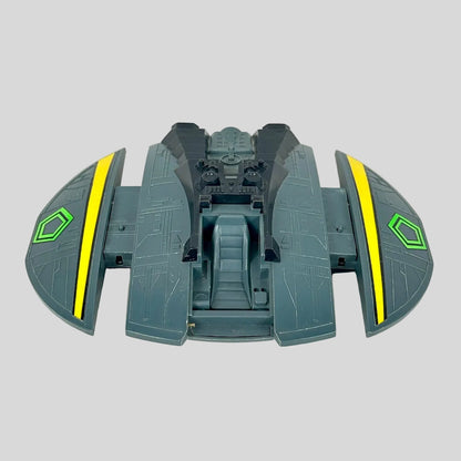A toy spaceship accessory in dark gray-green with bright yellow stripes and a green hexagonal emblem.