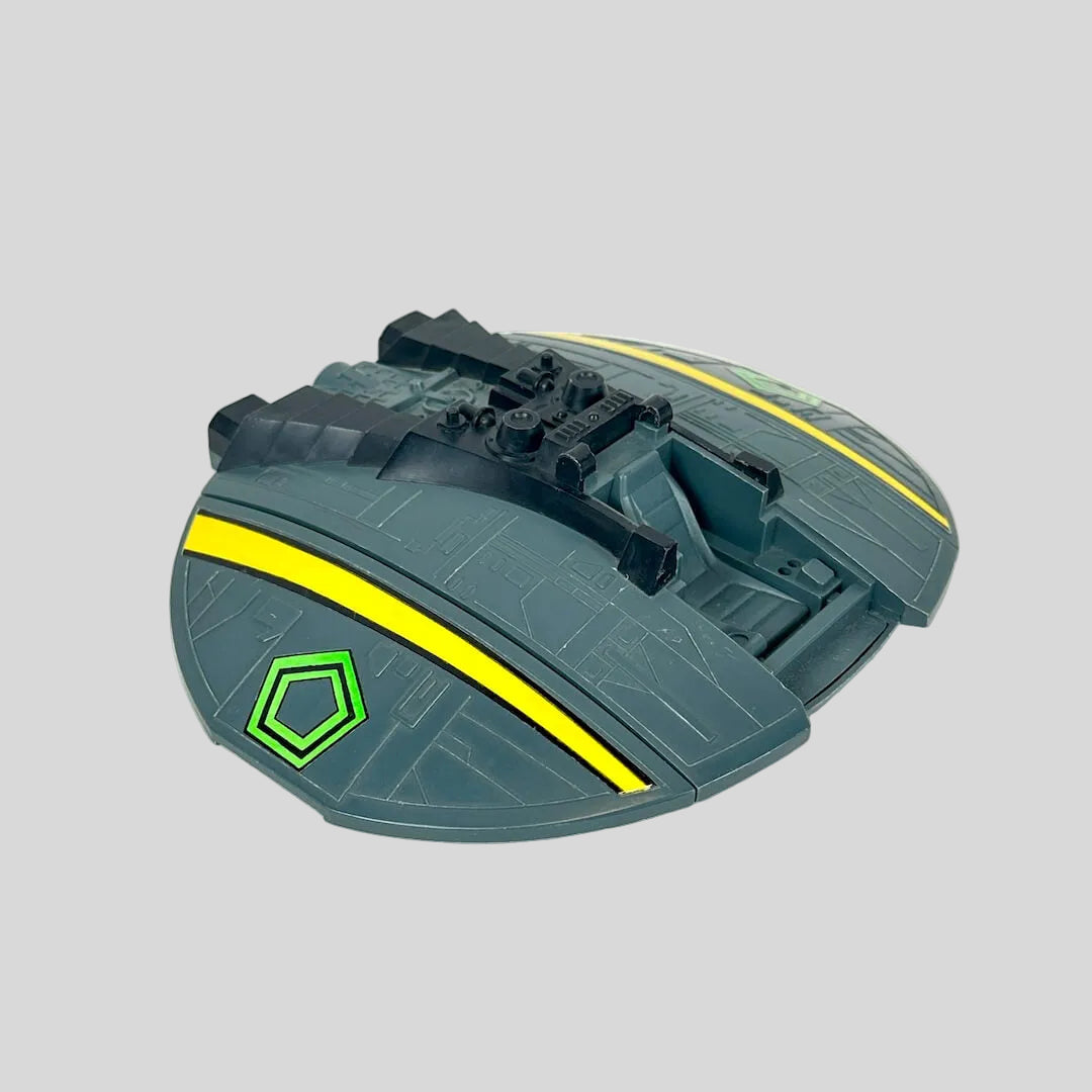 A toy spaceship accessory in dark gray-green with bright yellow stripes and a green hexagonal emblem.