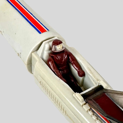 A vintage toy vehicle cockpit with a small maroon astronaut figure seated inside, photographed on a light surface.