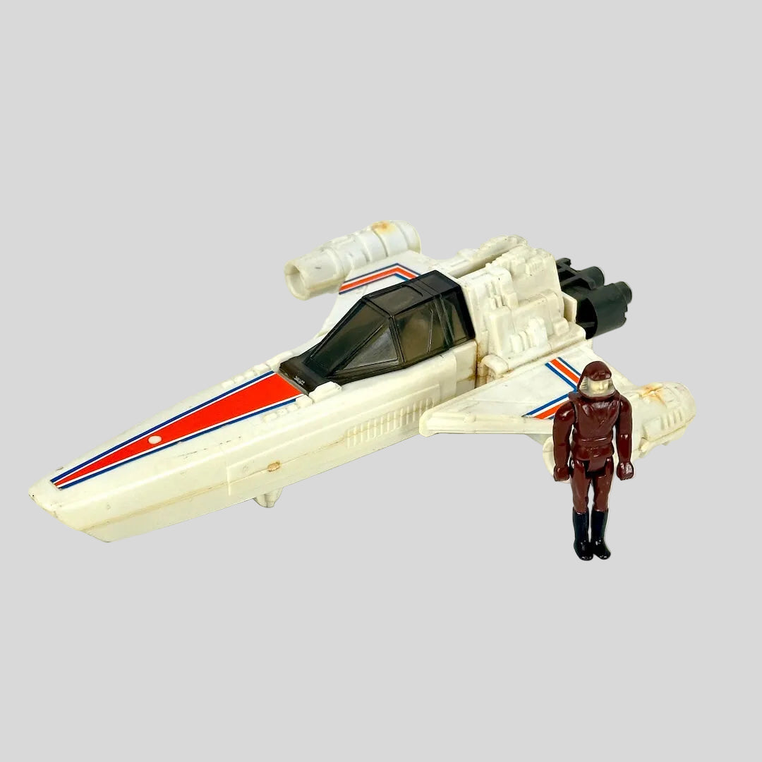 A vintage toy spacecraft in off-white with orange and blue stripes, shown next to a small brown astronaut figure. 