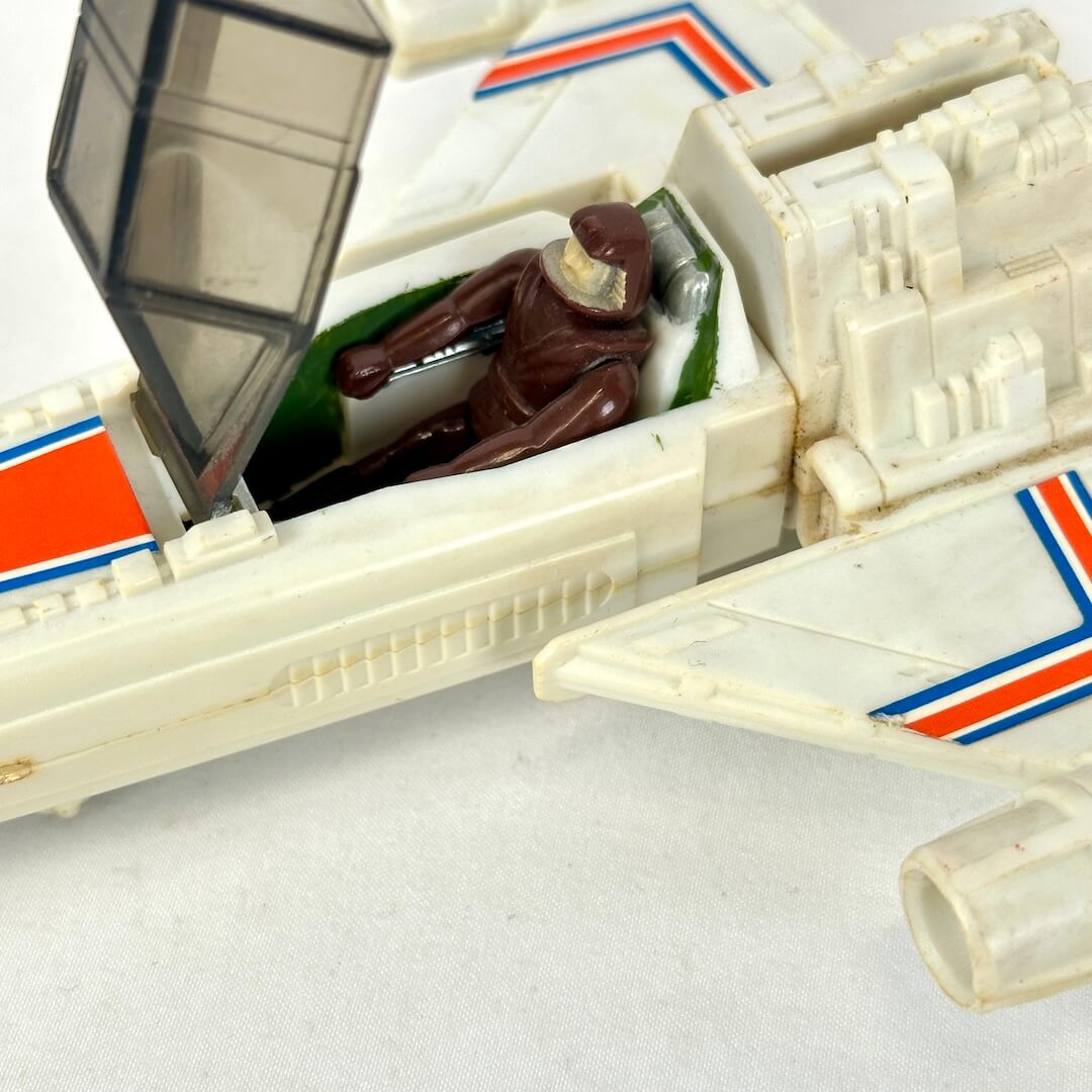 Vintage toy model of a white fighter jet with orange and blue stripes, featuring a small brown astronaut figure sitting in the cockpit.