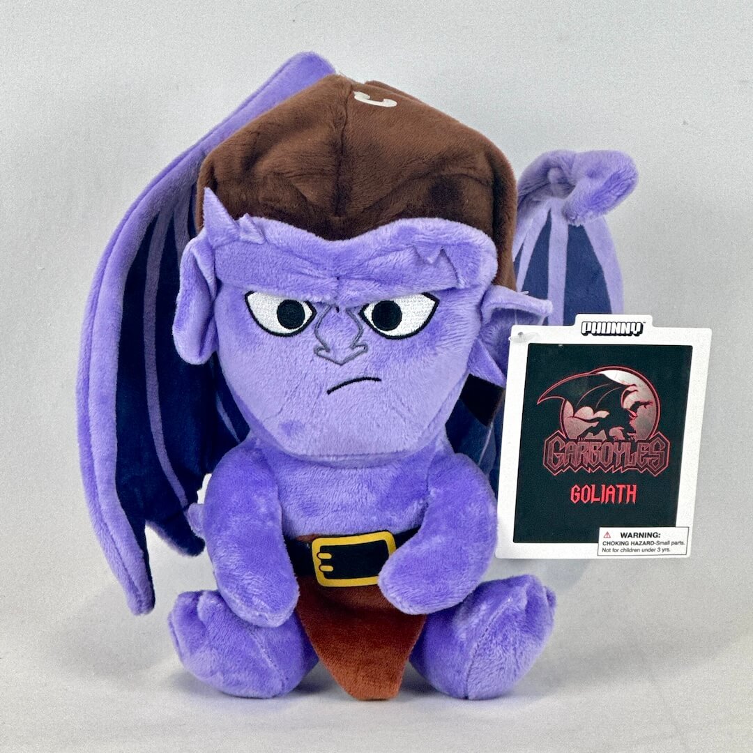 Purple cartoon character plush toy with a brown hat and pants, standing next to a card with 'Gargoyles' logo.