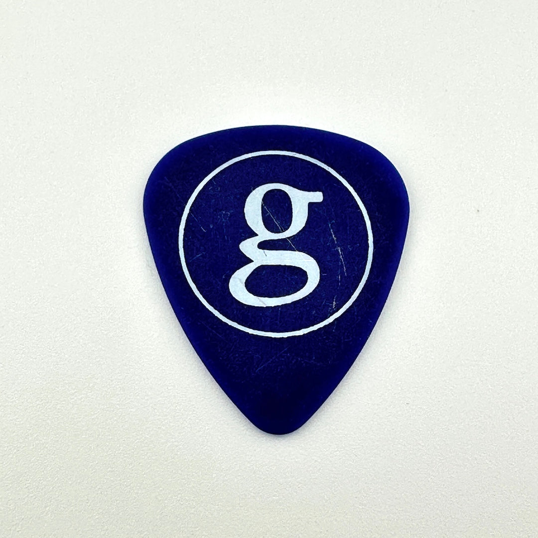 A dark blue guitar pick with a white circular logo containing a stylized lowercase 'g' in the center.