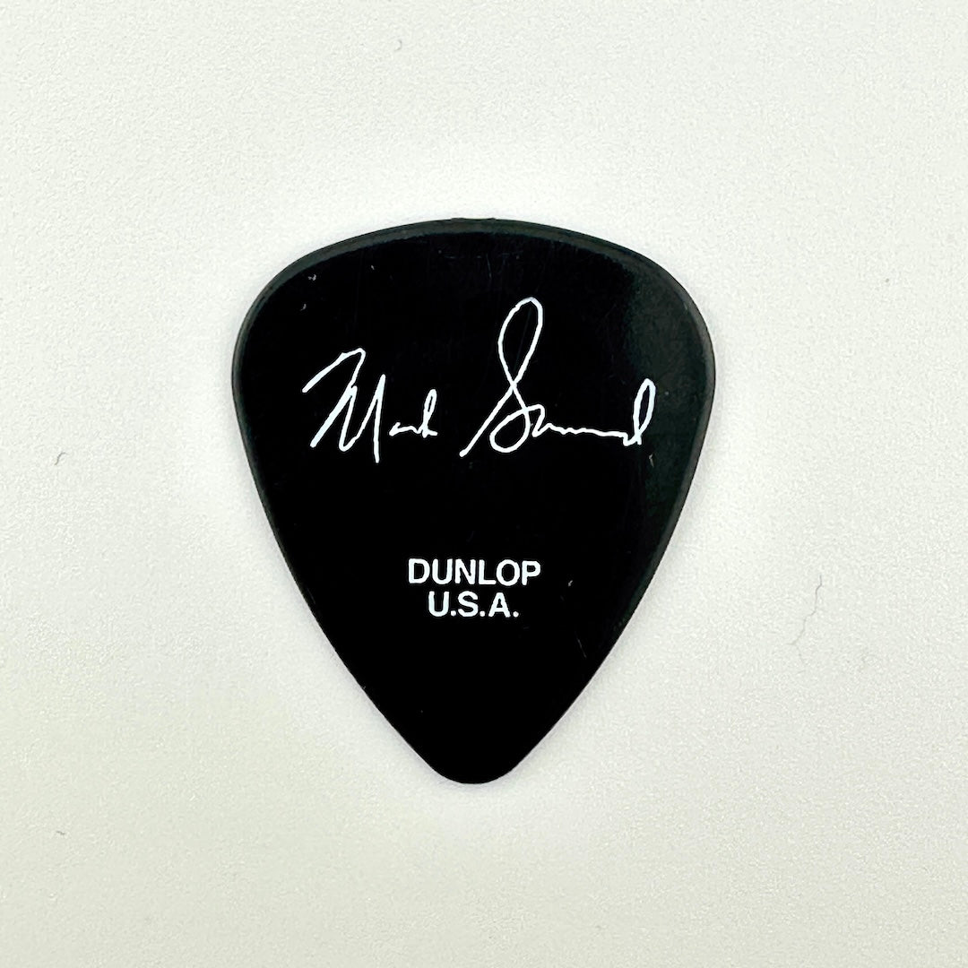 A black plastic guitar pick featuring a white autographed signature and the text 'DUNLOP U.S.