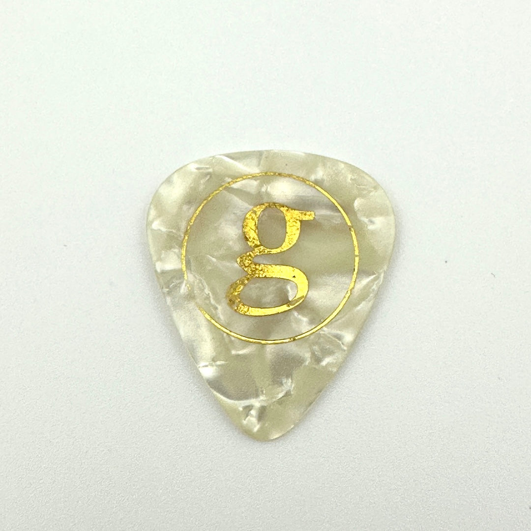 A single pearlescent translucent guitar pick with a gold glittery letter 'g' inside a gold circle, centered on a light gray background.
