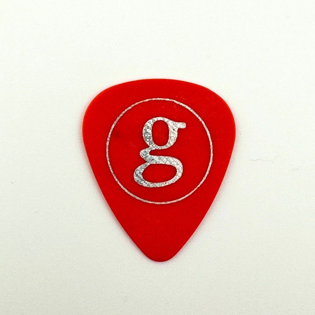 A red guitar pick featuring a silver glittery lowercase 'g' logo inside a thin silver circle, set against a plain off-white background.