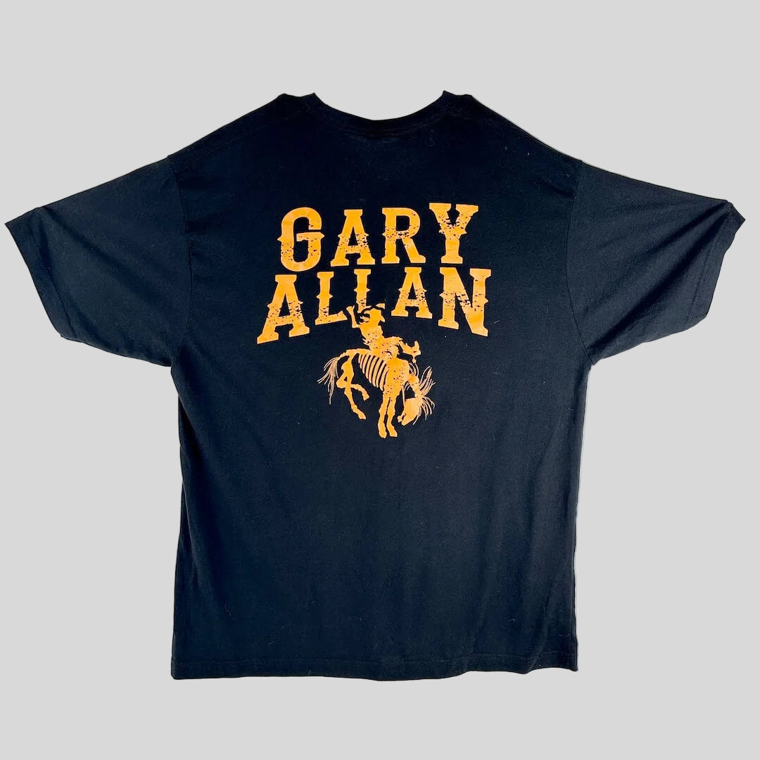 A black short-sleeve t-shirt laid, featuring a distressed yellow graphic that reads 'GARY ALLAN' above a cowboy riding a bucking horse.