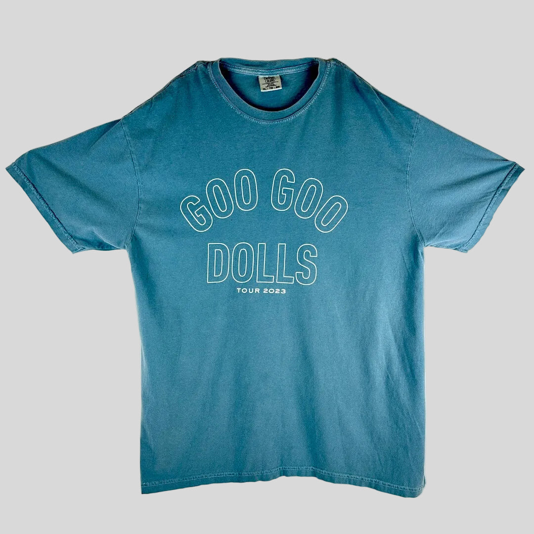 A faded blue short-sleeve t-shirt laid flat, featuring the text 'GOO GOO DOLLS' and 'TOUR 2023' printed in white outline on the front.