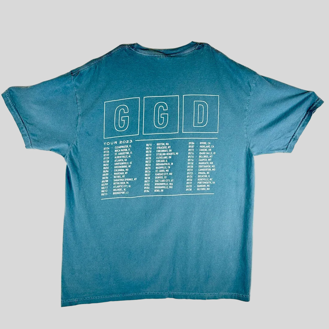 A blue short-sleeve t-shirt laid, showing the back side with a white graphic that reads 'GGD' in three boxes above a tour dates list.