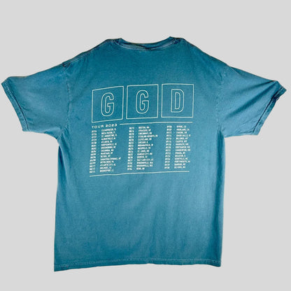 A blue short-sleeve t-shirt laid, showing the back side with a white graphic that reads 'GGD' in three boxes above a tour dates list.