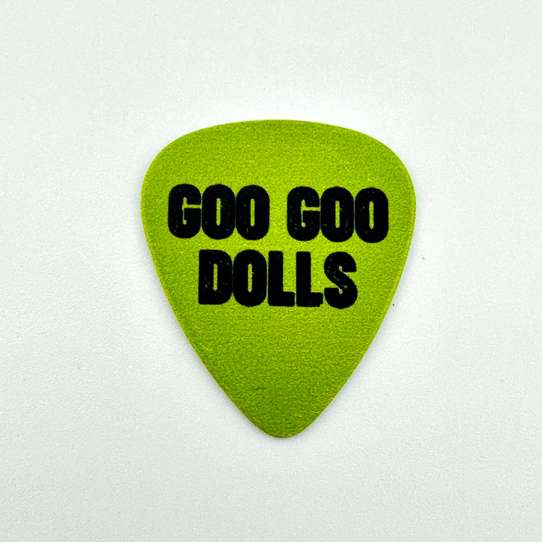 A lime green guitar pick with the text 'GOO GOO DOLLS' printed in bold black letters, centered on a light off-white background.