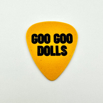 A yellow guitar pick with the text 'GOO GOO DOLLS' printed in black, centered on a light gray background.