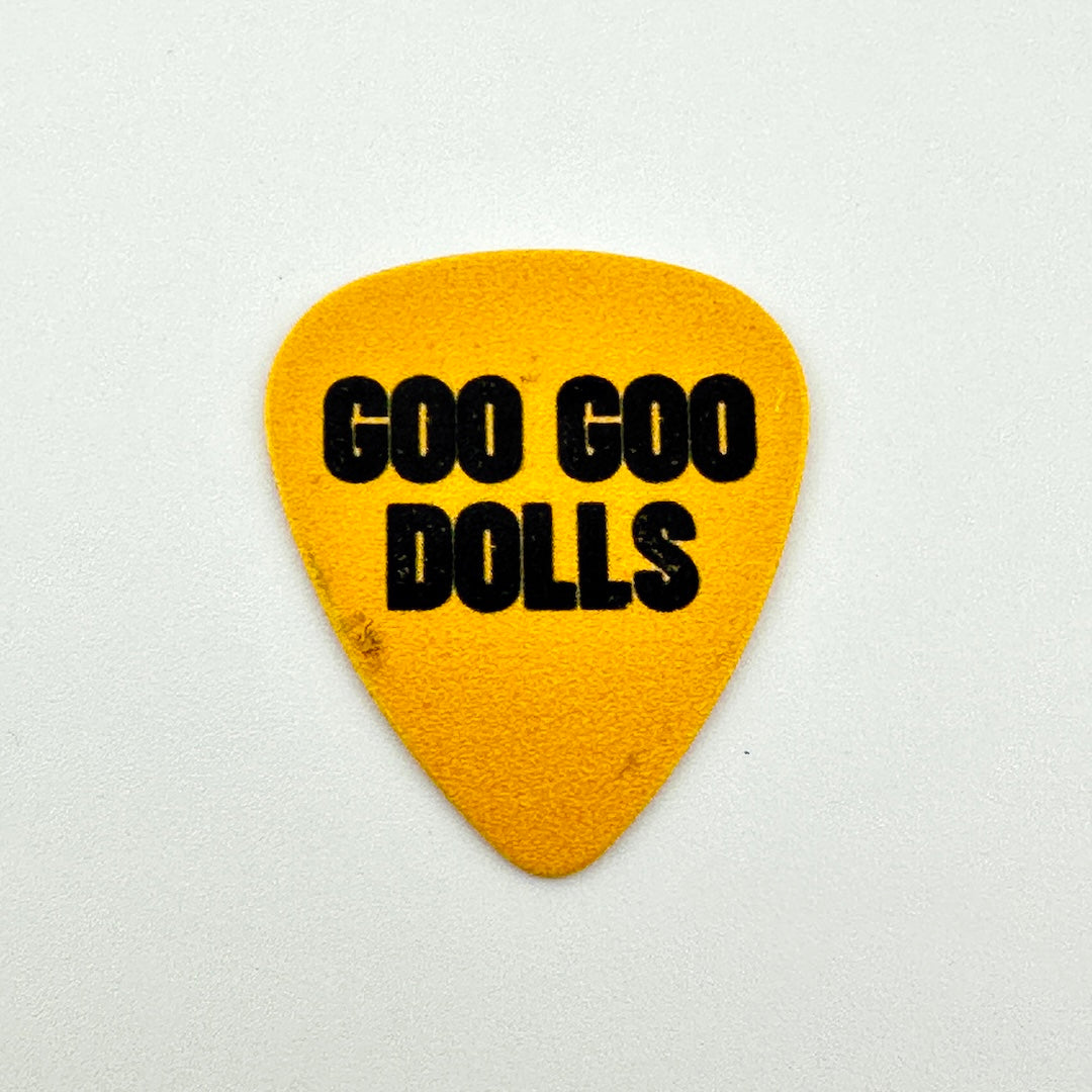A yellow guitar pick with the text 'GOO GOO DOLLS' printed in bold black letters, centered on a light gray background.