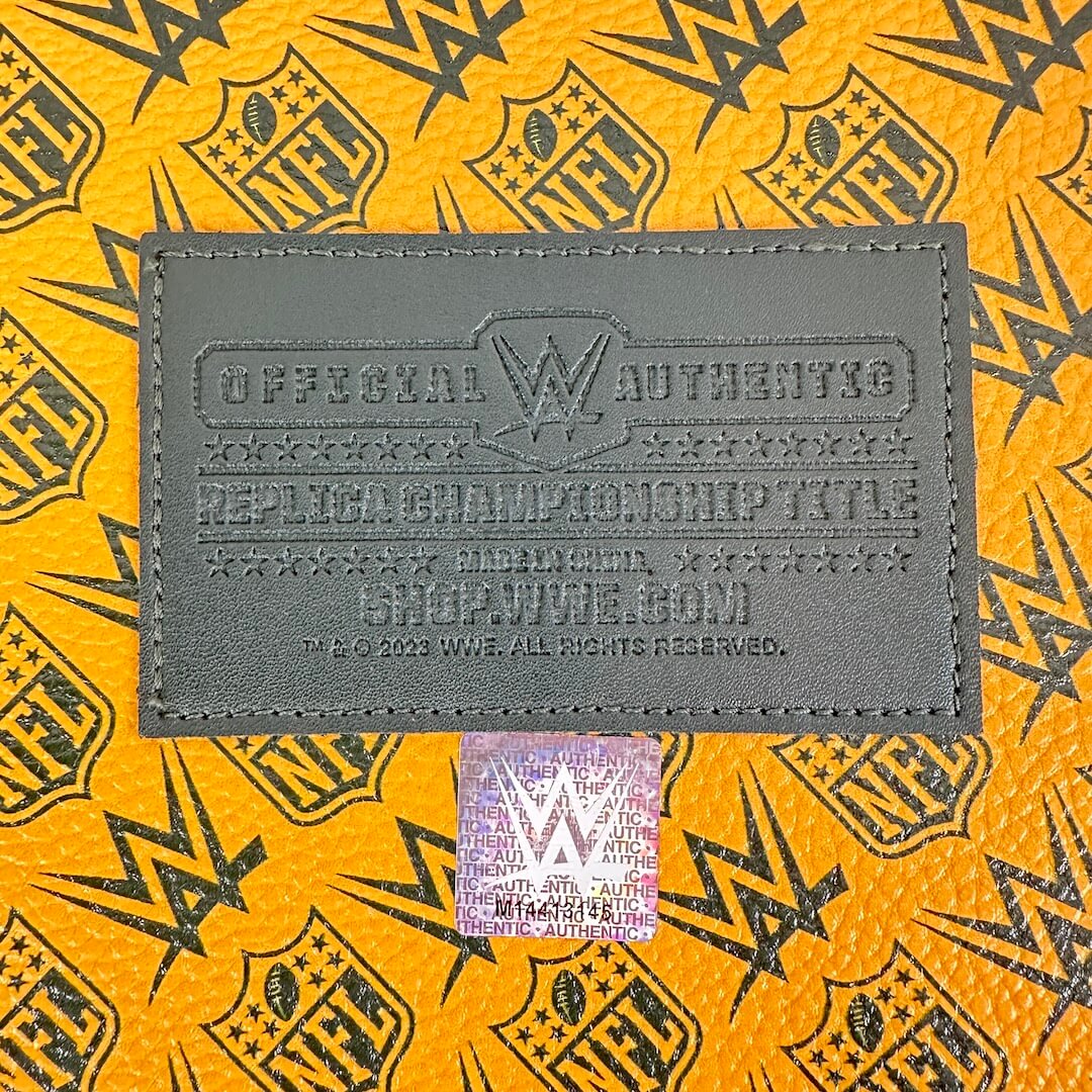 A close-up of a dark gray WWE official authenticating patch on a bright yellow and black patterned surface, with a holographic WWE sticker below it.