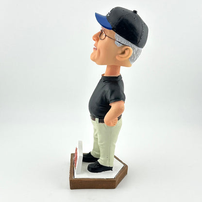 Bobblehead figure of a man wearing a black shirt, beige pants, and a blue and black cap on a white background.