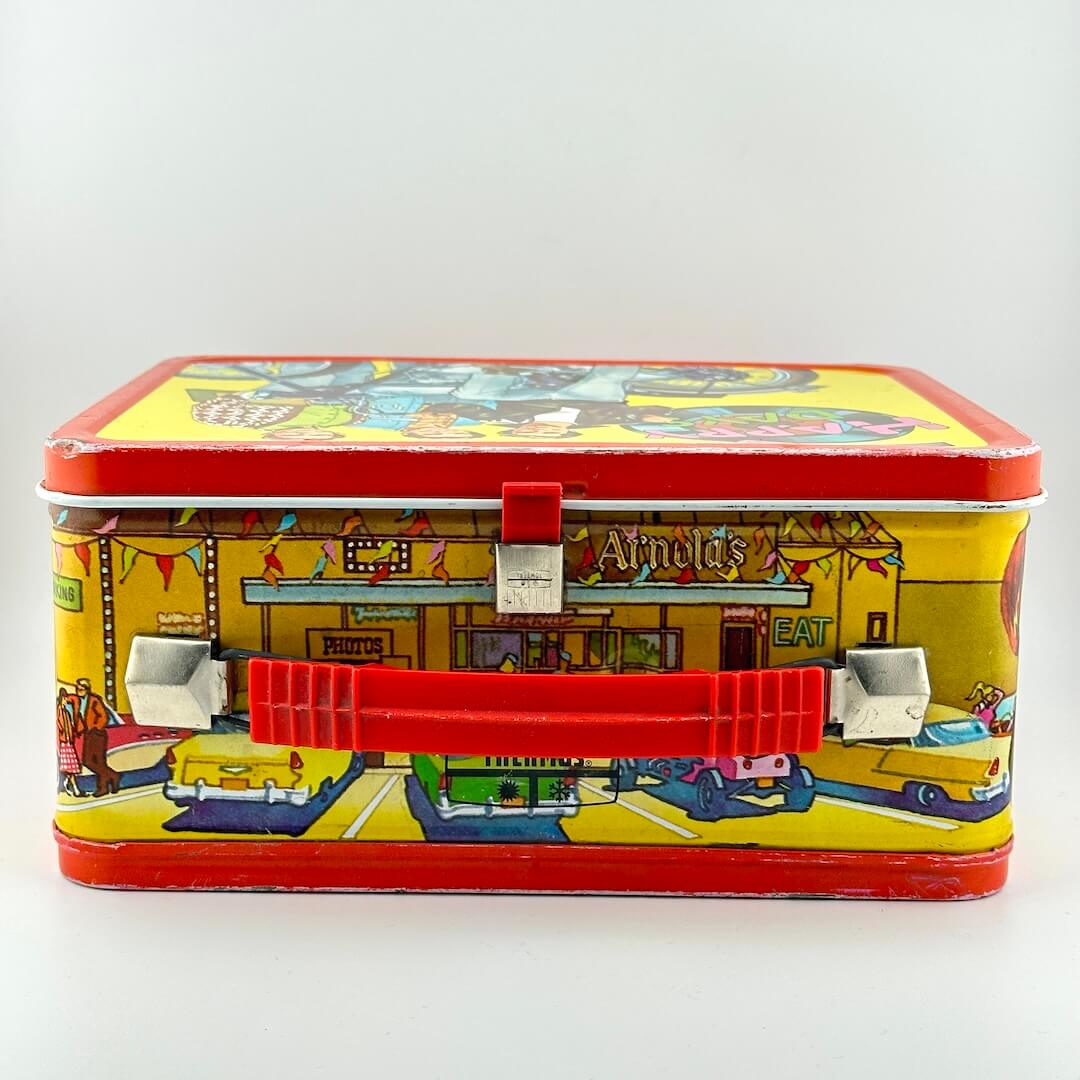 Top view of metal lunchbox showing red plastic latch and handle, red border and yellow background. 