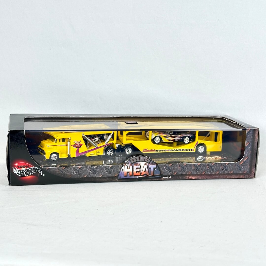 Hot Wheels toy set featuring a yellow race car transporter truck carrying two diecast race cars in their original packaging. 