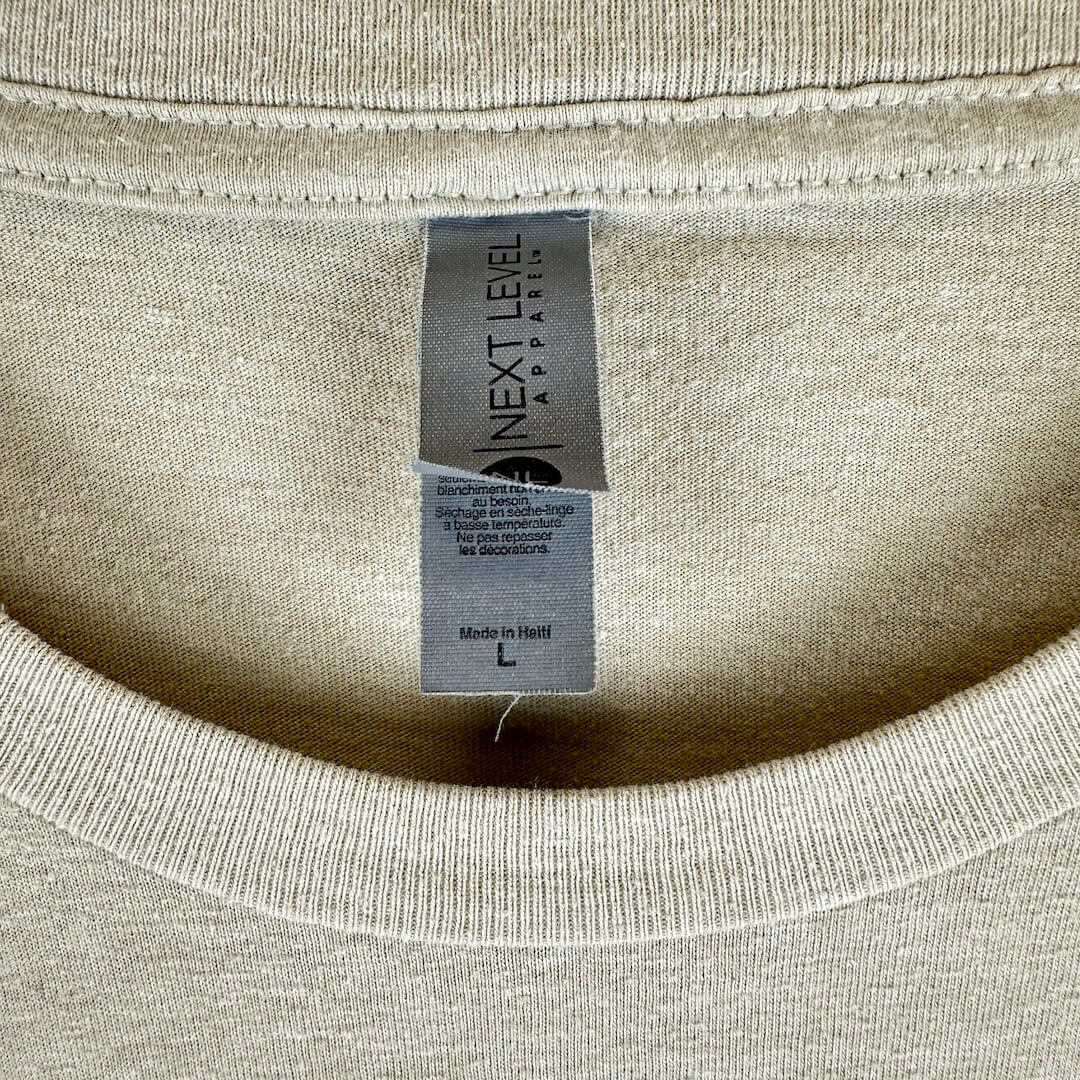 Close-up of the inside neck of a light beige crewneck t-shirt, showing the brand label 'NEXT LEVEL APPAREL' & size information.