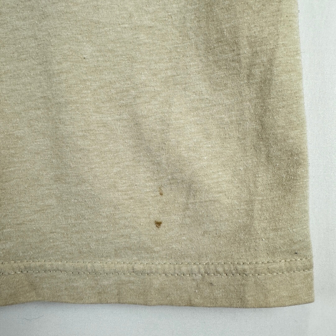 A close-up of a light beige heathered fabric, showing a hemmed edge and a small brown stain on the surface.
