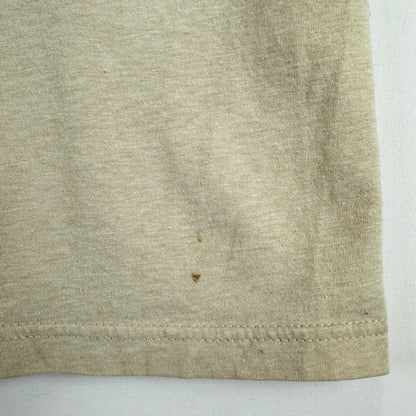 A close-up of a light beige heathered fabric, showing a hemmed edge and a small brown stain on the surface.