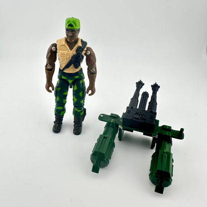 Toy figure with green helmet and camouflage pants, standing next to a green and black weapon on a white background