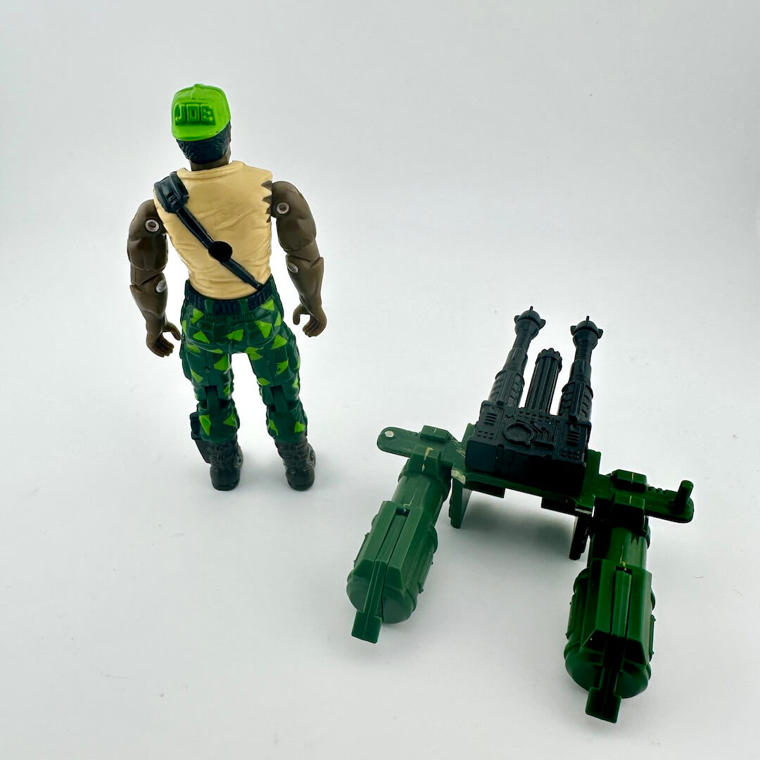 Toy figure with green helmet and camouflage pants, standing next to a green and black weapon on a white background